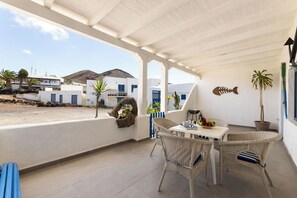 A covered outdoor seating area with wicker furniture, a table with a fruit bowl, and a view of the surrounding buildings and landscape. - Home2Book Casa Yoyo Lanzarote Coastal Home (El Golfo)