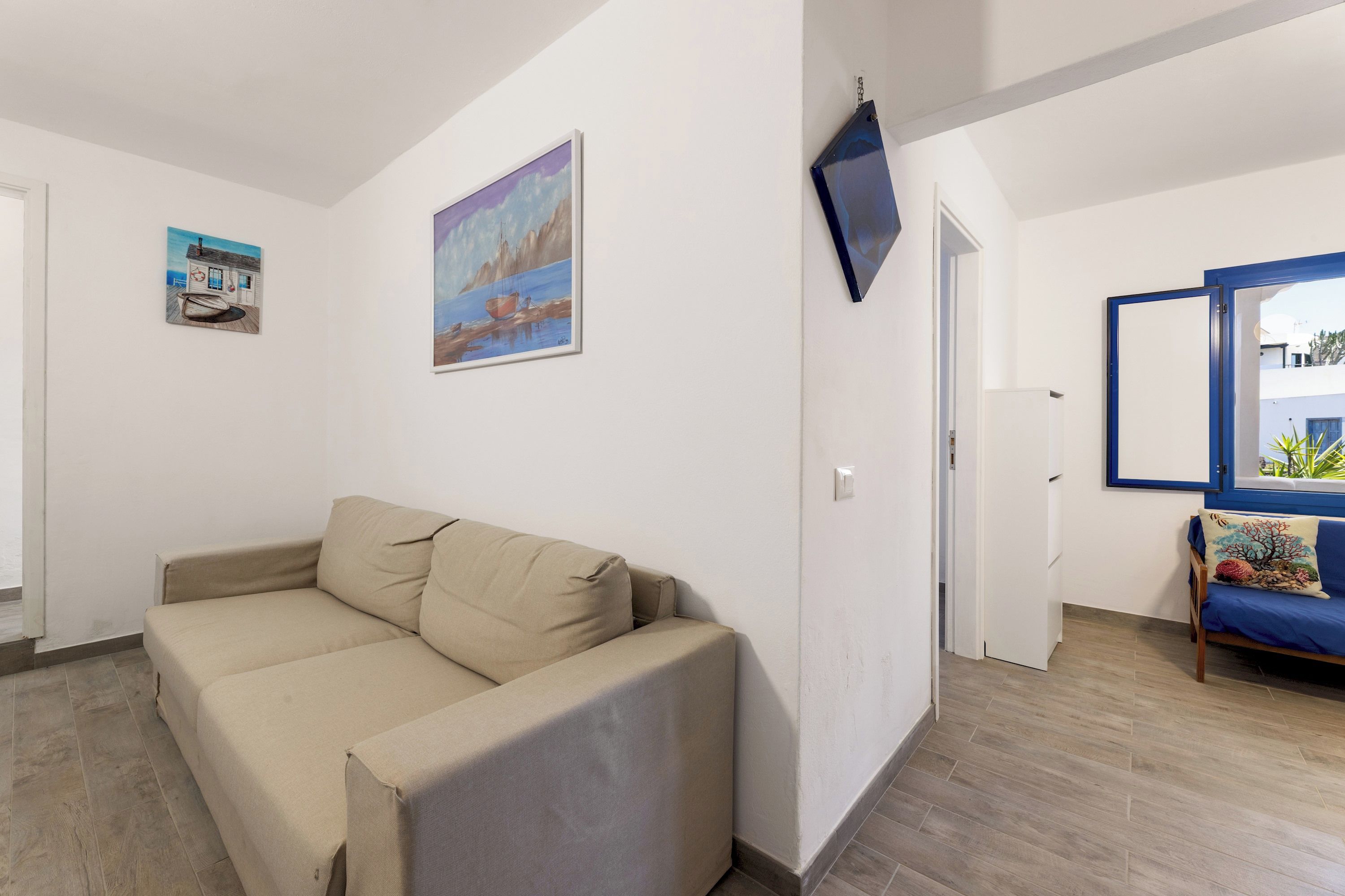 Una sala de estar con un sofá color beige, un cuadro que muestra un barco en el agua, y un espejo con marco azul que refleja una vista de un edificio y plantas.