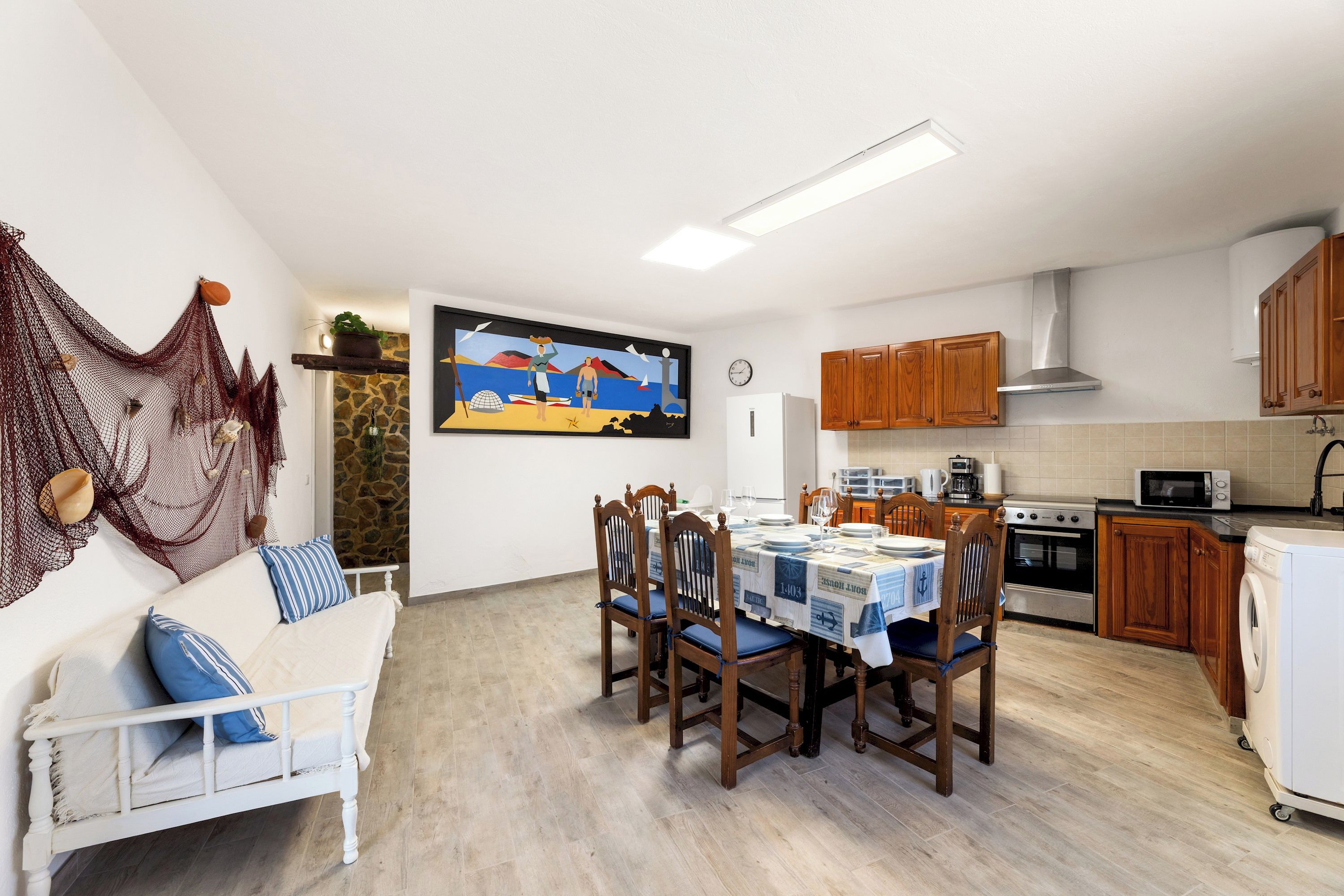 A dining area with a table set for four, wooden chairs, a white sofa with blue cushions, a painting on the wall, and a kitchen with wooden cabinets and appliances.