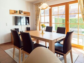 A dining area with a wooden table, black chairs, a TV mounted on the wall, and a window with curtains. - 6b/t5 Holiday Park "freesenbruch" (Zingst)