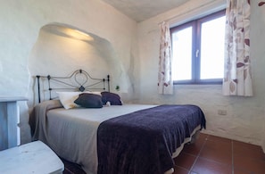 A bedroom with a bed, a bedside table, a window with curtains, and a wall with a unique texture. - Artenara Retreat With Hot Tub (Artenara)