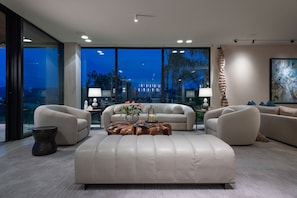 A modern living room with a large sectional sofa, a coffee table, and a view of a cityscape at dusk. - Casa Mare Luxury Estates Rentals At Kupuri (Punta Mita)