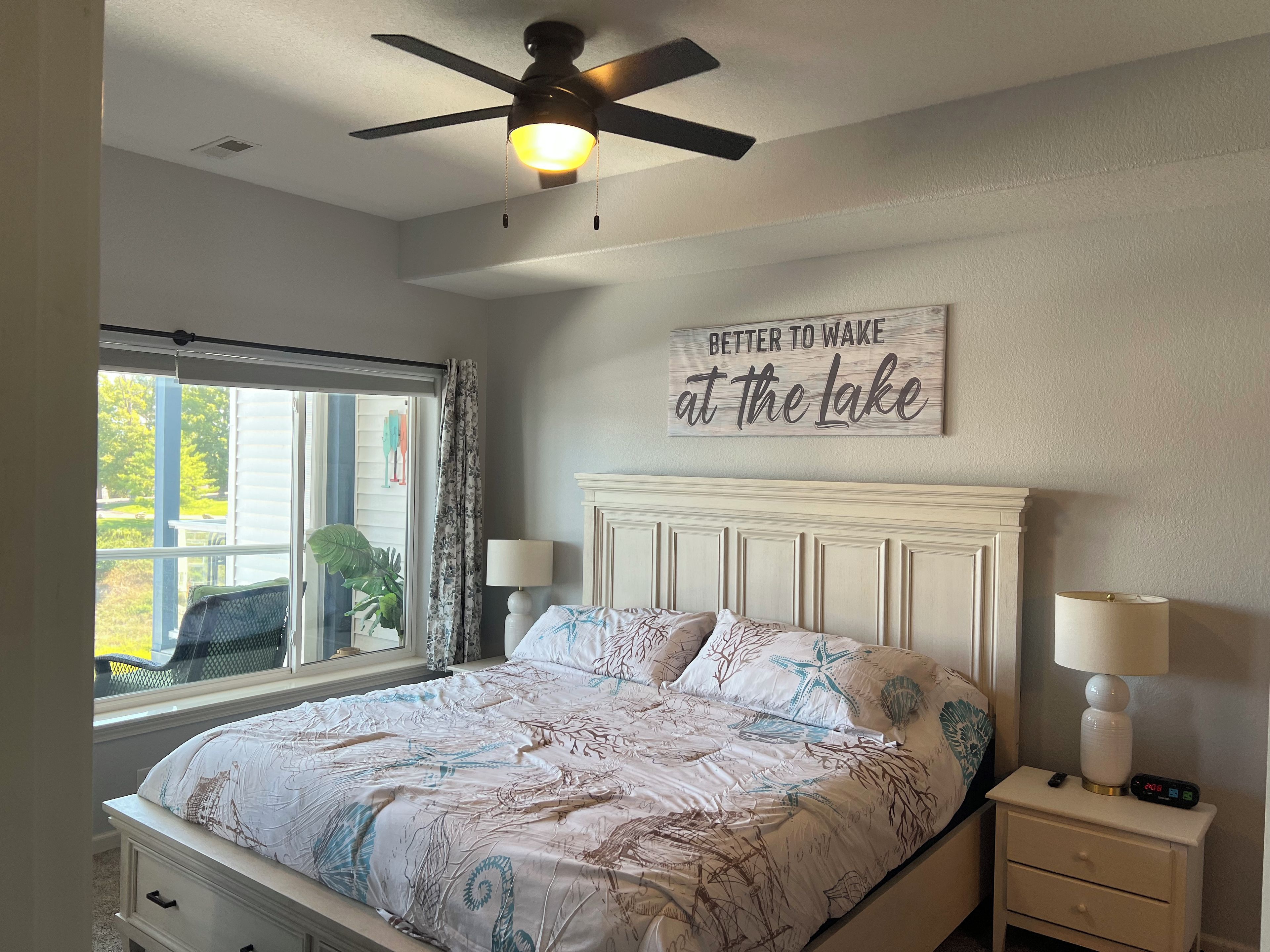Une chambre à coucher comprenant un lit, une table de chevet, une lampe, un ventilateur de plafond, une fenêtre avec des rideaux et un panneau décoratif mural.