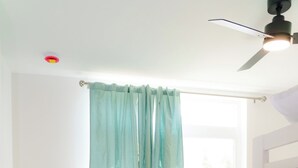 Una cama individual con colcha verde, una ventana con cortinas azul claro y un ventilador de techo.