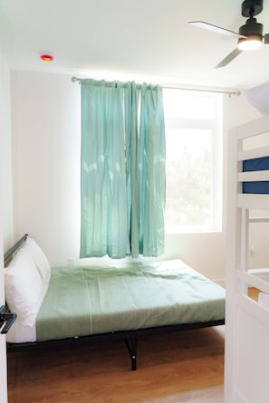 A single bed with a green bedspread, a window with light blue curtains, and a ceiling fan. - Coastal Elegance - 1 Block to Beach - EV Charging - Parking (Seaside Heights)