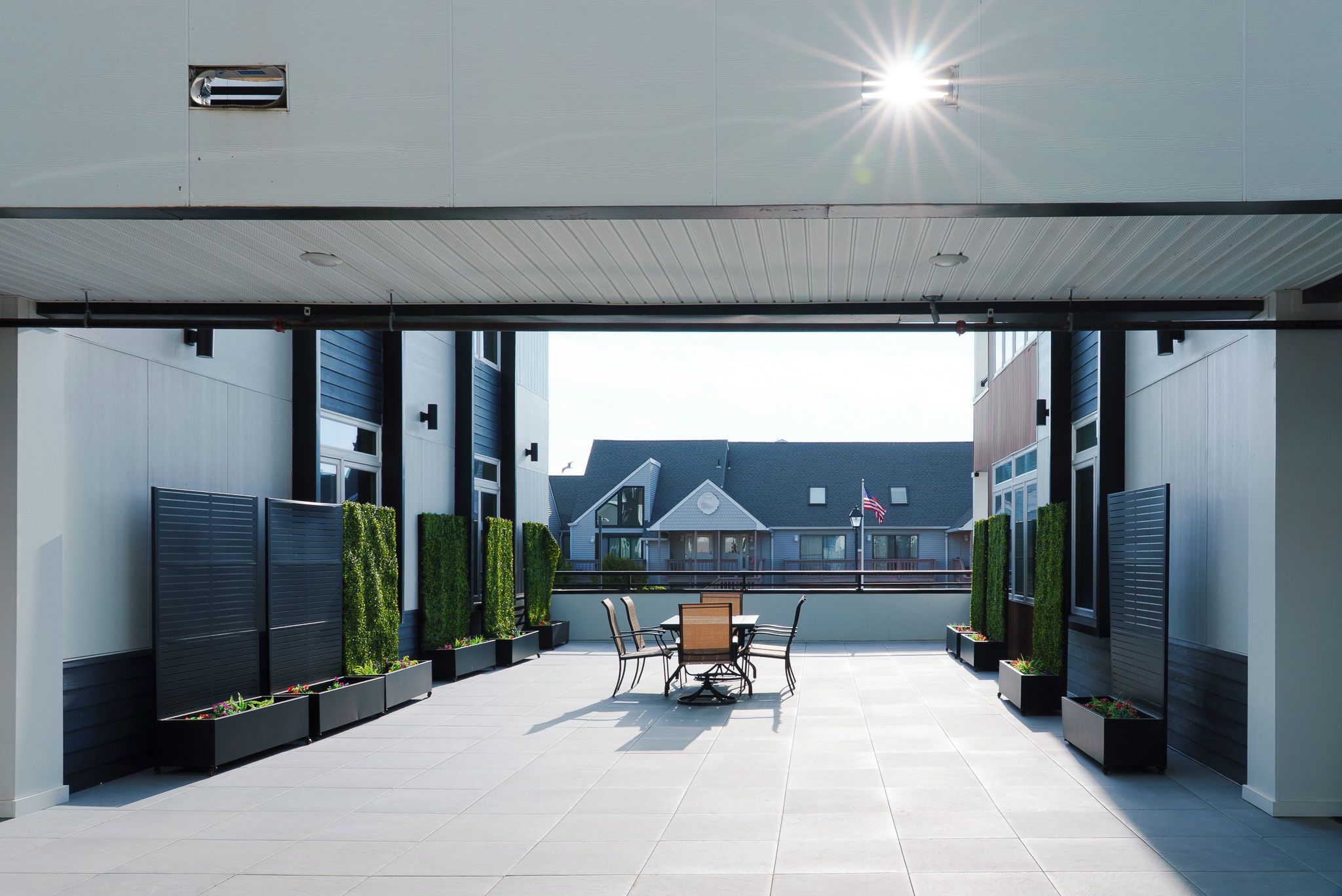 A covered outdoor patio with a table and chairs, surrounded by planters and a view of a residential building.