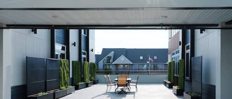 A covered outdoor patio with a table and chairs, surrounded by planters and a view of a residential building.