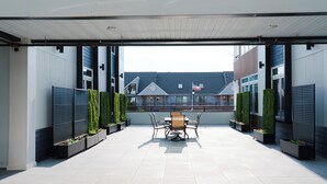 Un patio al aire libre con una mesa y sillas, rodeado de macetas con follaje, y vistas a edificios residenciales al fondo.