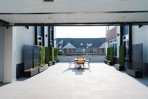 An outdoor patio area with a table and chairs, surrounded by planters with greenery, and a view of residential buildings in the background. - Coastal Elegance - 1 Block to Beach - EV Charging - Parking (Seaside Heights)