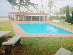 A rectangular swimming pool with a brick border, surrounded by a grassy area and a building with a tiled roof.