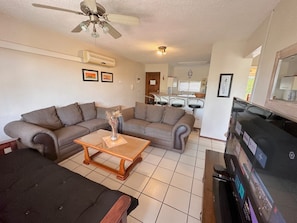A living room with a sofa, a coffee table, and a TV. There is a ceiling fan and an air conditioning unit.