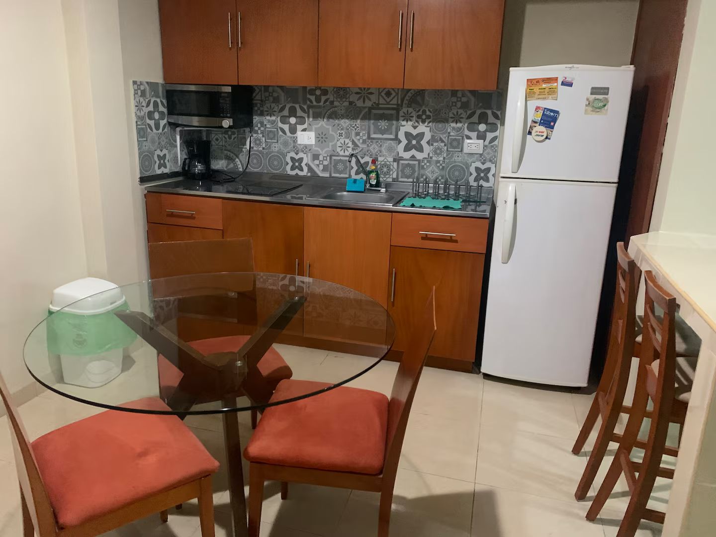 A compact kitchen with wooden cabinets, a white refrigerator, a glass table, and red cushioned chairs.