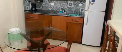 A compact kitchen with wooden cabinets, a white refrigerator, a glass table, and red cushioned chairs.