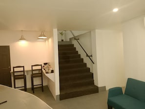 A staircase leading to an upper floor, a dining area with chairs and a table, and a teal sofa in the living area.