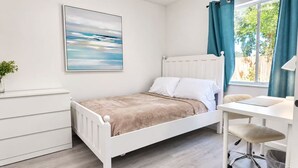 A bedroom with a bed, a desk, a chair, a radiator, a window with curtains, and a painting on the wall.