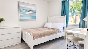 A bedroom with a bed, a desk, a chair, a radiator, a window with curtains, and a painting on the wall.
