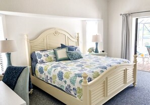 A bed with a floral bedspread, a bedside table with a lamp, and a view of the outdoors through a window.