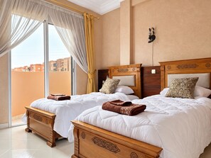 Quarto de hotel com duas camas, uma janela grande com cortinas, vista de edifícios e luminária fixada na parede.
