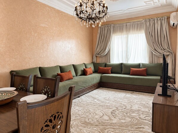 Uma sala de estar com um sofá seccional, uma mesa de jantar com cadeiras, um lustre e uma janela grande com cortinas.