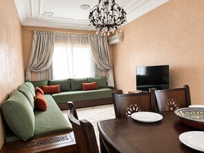 Uma sala de estar com um sofá verde, uma mesa de jantar com cadeiras, uma televisão e um candelabro.