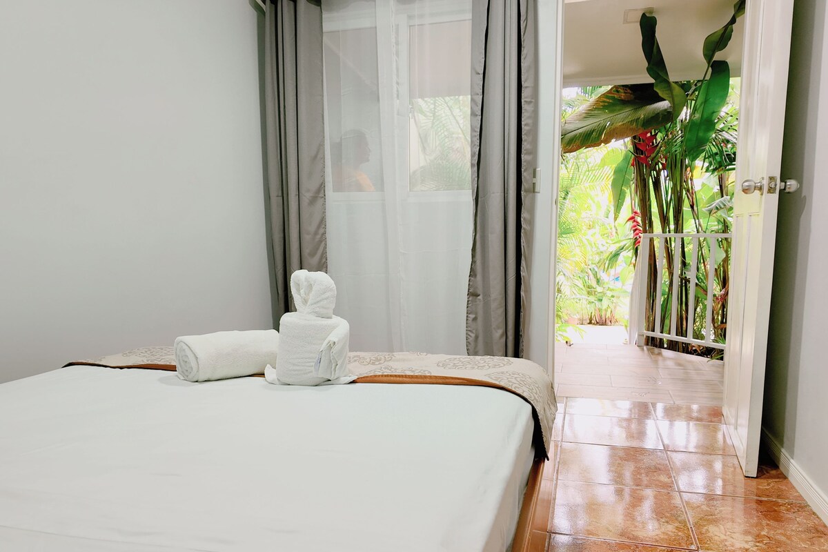 A hotel room with a bed, a bathroom with a shower curtain, and a view of a garden through an open door.