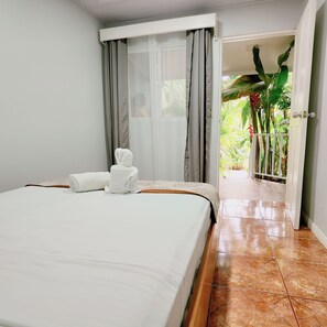 A hotel room with a bed, a bathroom with a shower curtain, and a view of a garden through an open door. - Toucan Arenal Lodge (La Fortuna)