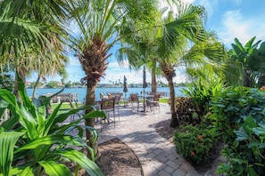 Une terrasse extérieure avec des tables et des chaises, entourée de palmiers et de plantes, donnant sur une étendue d’eau.
