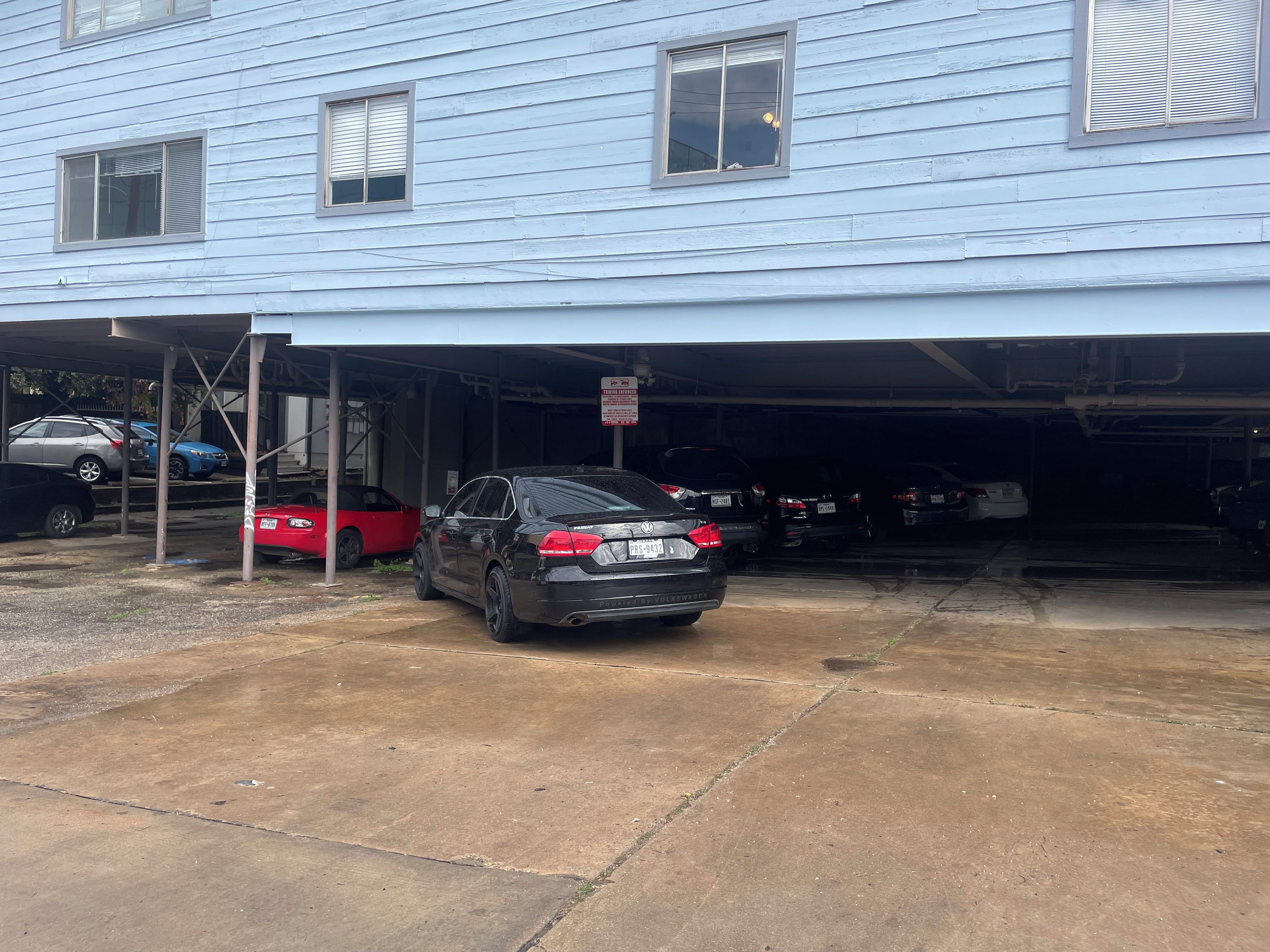 A garage with a black car parked inside, a red car parked outside, and a blue building in the background.