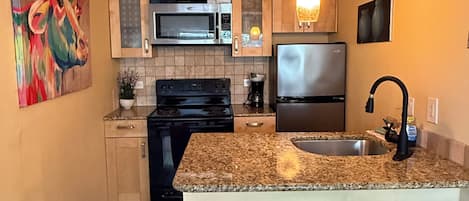 A kitchen with wooden cabinets, a microwave, stove, refrigerator, granite countertop, and two bar stools.
