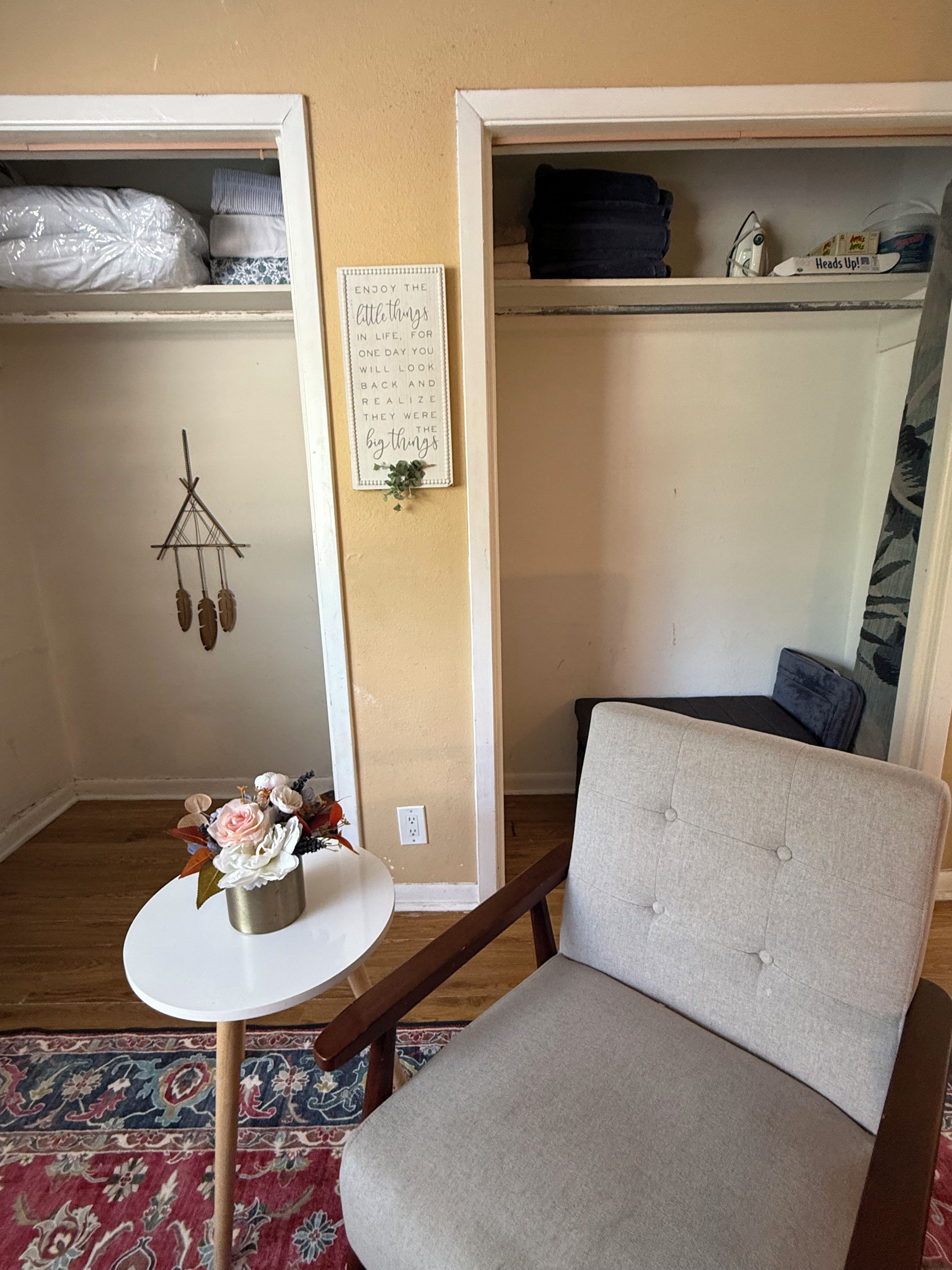 A room with a grey upholstered armchair, a small round table with a floral arrangement, a wall-mounted decorative item, and a patterned rug.