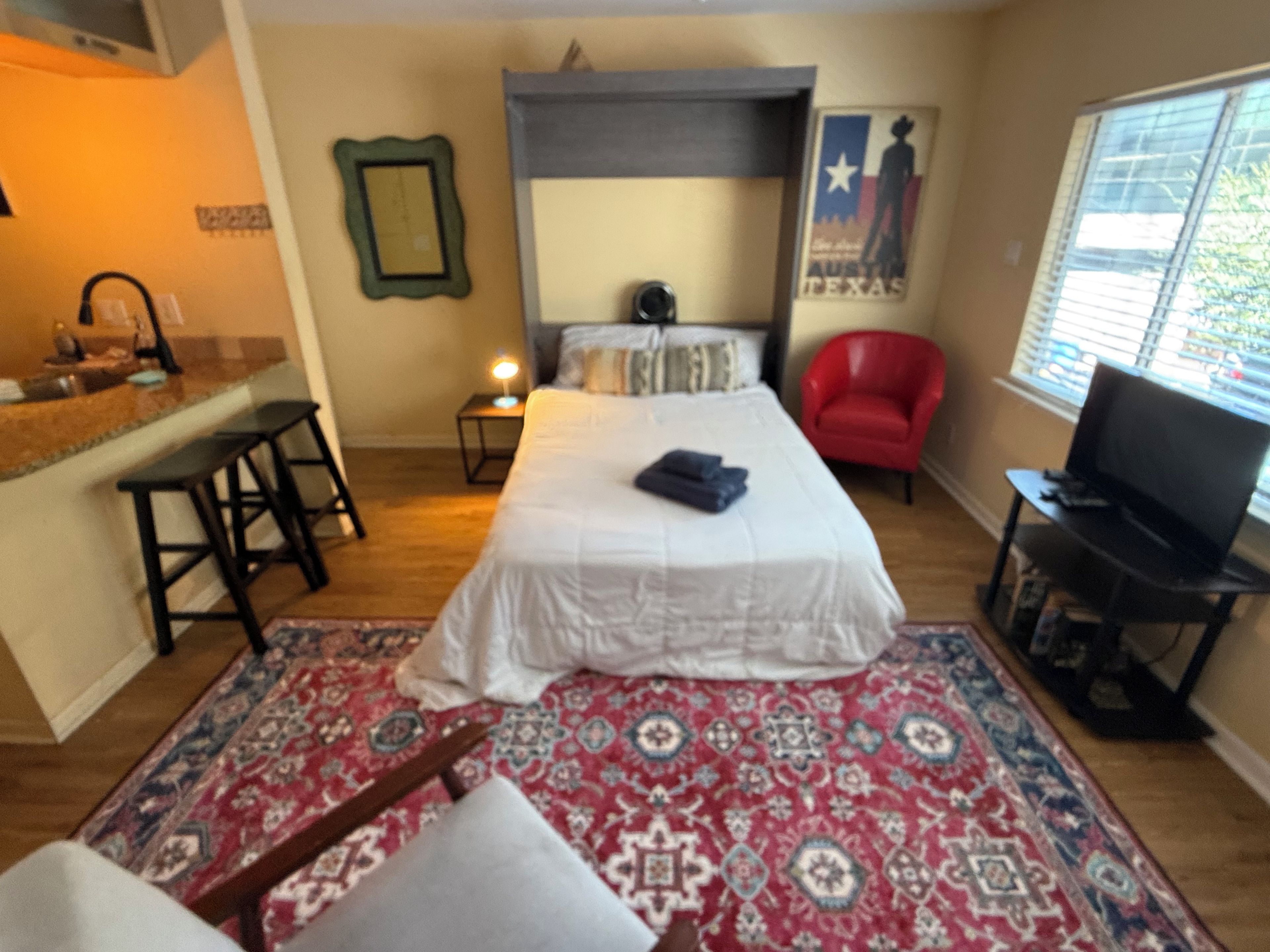 A hotel room with a bed, a desk, a red chair, a television, and a window with blinds.