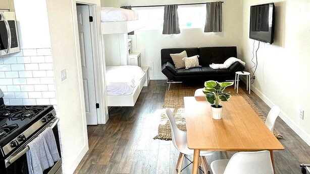 A compact living space with a kitchenette, a sofa, a dining table with chairs, and a TV mounted on the wall. - Cozy studio home - Near BYU and UVU (Orem)