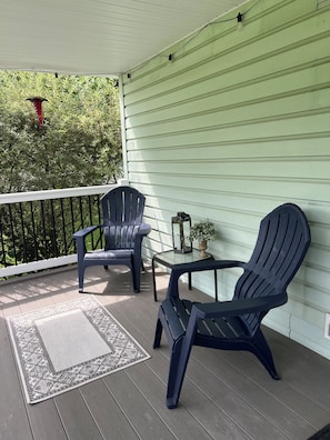 Varanda coberta com duas cadeiras Adirondack, uma mesinha com uma planta e um candeeiro decorativo.
