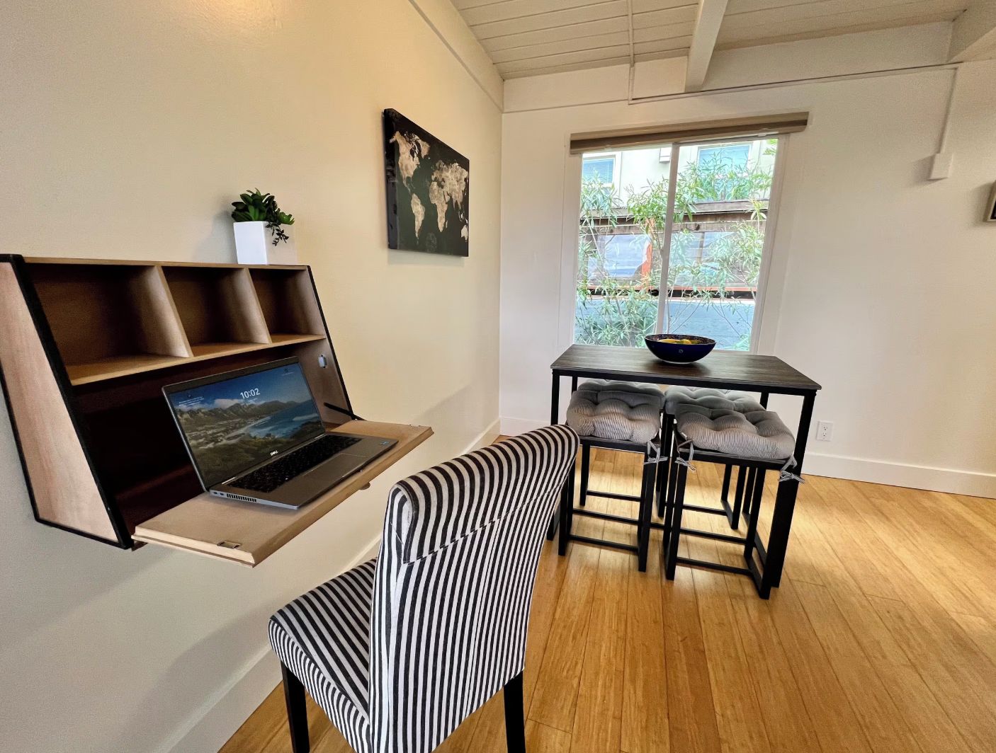 A modern room with a laptop on a shelf, a dining table with chairs, and a window with a view of greenery.
