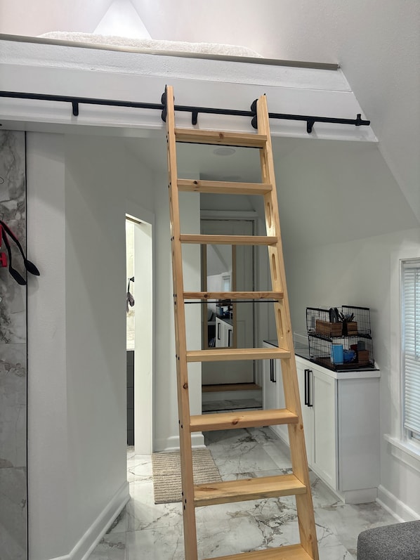 A wooden ladder against a white wall in a room with a marble floor, a black metal rack with items on top, and a door leading to another room.