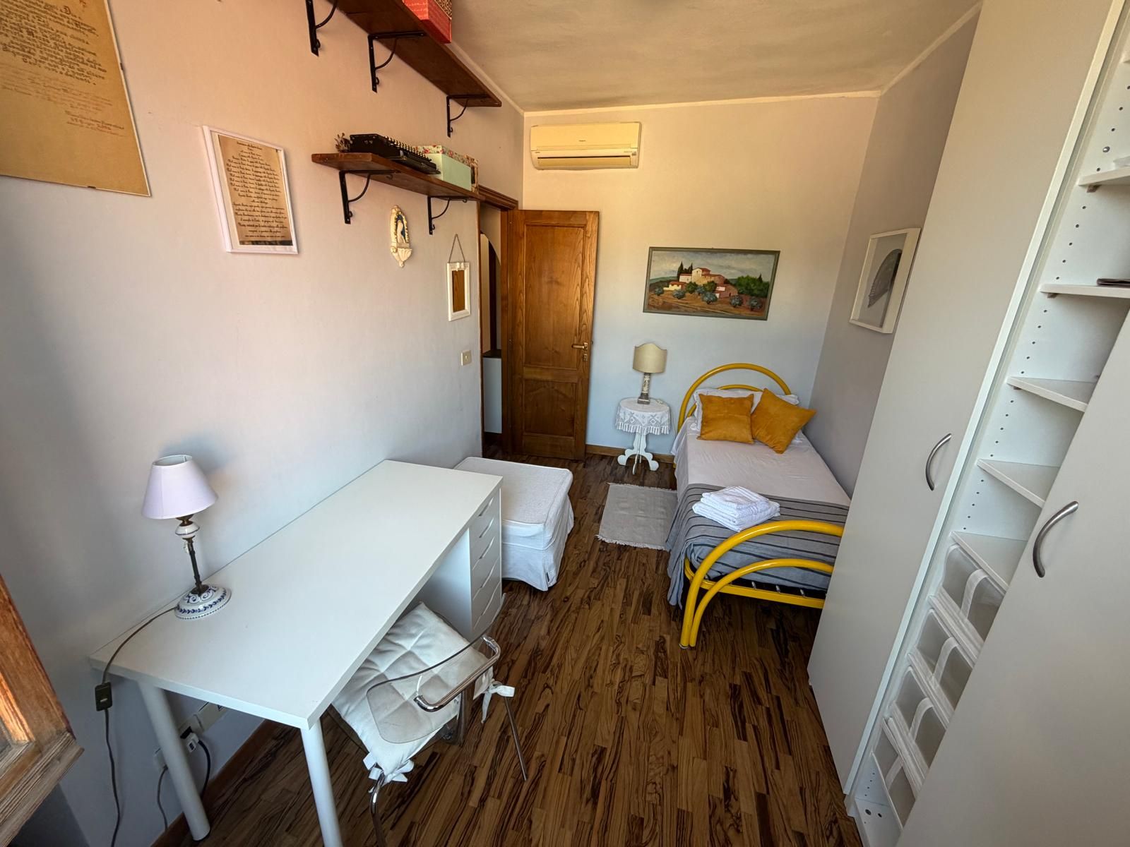 Ein kleines Zimmer mit einem Bett, einem Schreibtisch, einem Stuhl, einem kleinen Tisch und einem Regal mit Gegenständen.