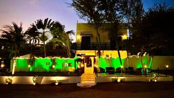 A building with a staircase leading to a door, surrounded by palm trees and illuminated by warm lights.