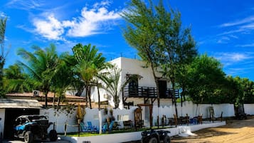A white building with a balcony, surrounded by palm trees and a sandy area with beach chairs.