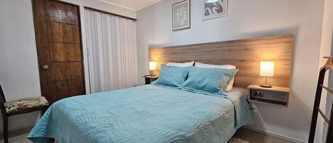 A bedroom with a bed, two bedside lamps, a wooden headboard, a door, and a window with curtains.