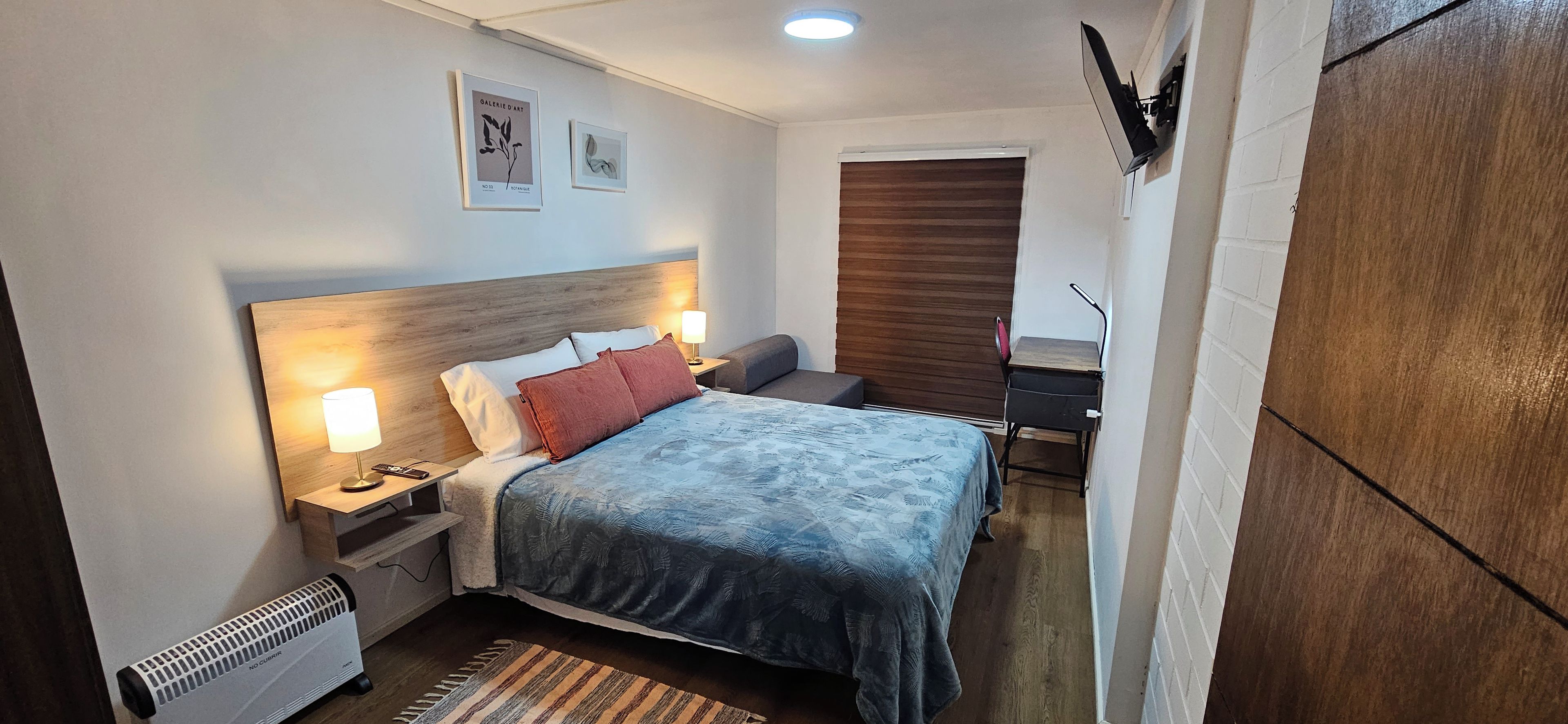 A hotel room with a bed, bedside lamps, a desk, a chair, and a radiator.