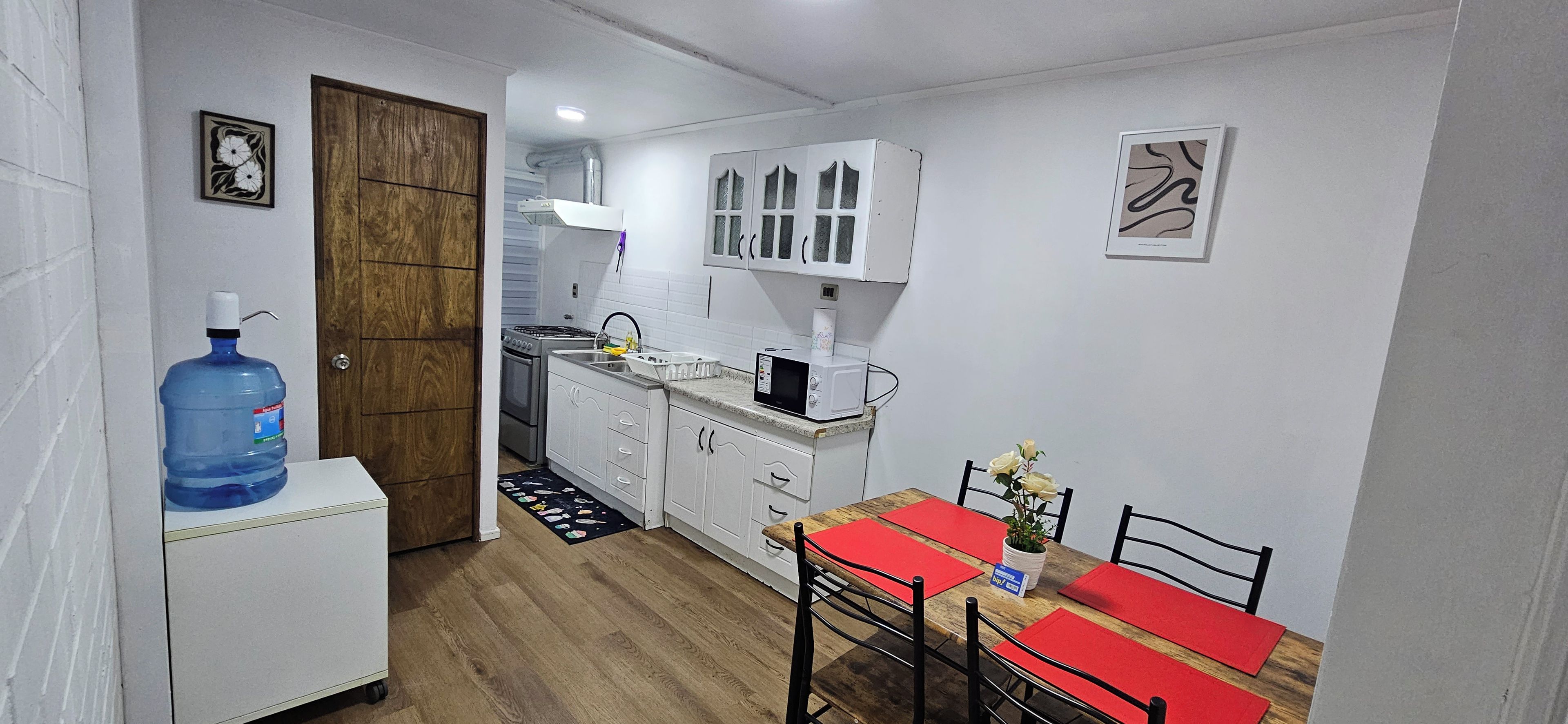 A compact kitchen with white cabinets, a microwave, and a sink. There's a large water dispenser and a dining table with red placemats.