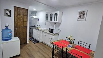 A compact kitchen with white cabinets, a microwave, and a sink. There's a large water dispenser and a dining table with red placemats.
