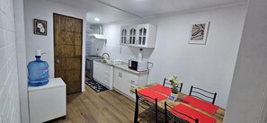 Une cuisine compacte avec des meubles blancs, un four à micro-ondes et un évier. On y trouve un grand distributeur d’eau et une table à manger avec des sets de table rouges.