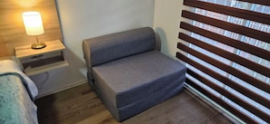 A small, compact room with a grey sofa, a bedside table with a lamp, and a window with blinds. - GUEST HOUSE AEROPUERTO PUDAHUEL (Santiago)