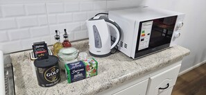 A kitchen counter with a microwave, a kettle, and various food items including a can of gold coffee, a box of Supremo herbal tea, and a jar of sugar.