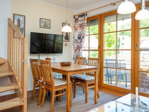 A dining area with a wooden table and chairs, a television mounted on the wall, and a view of the outdoors through large windows. - 4.5/t3 in the "freesenbruch" Holiday Park (Zingst)