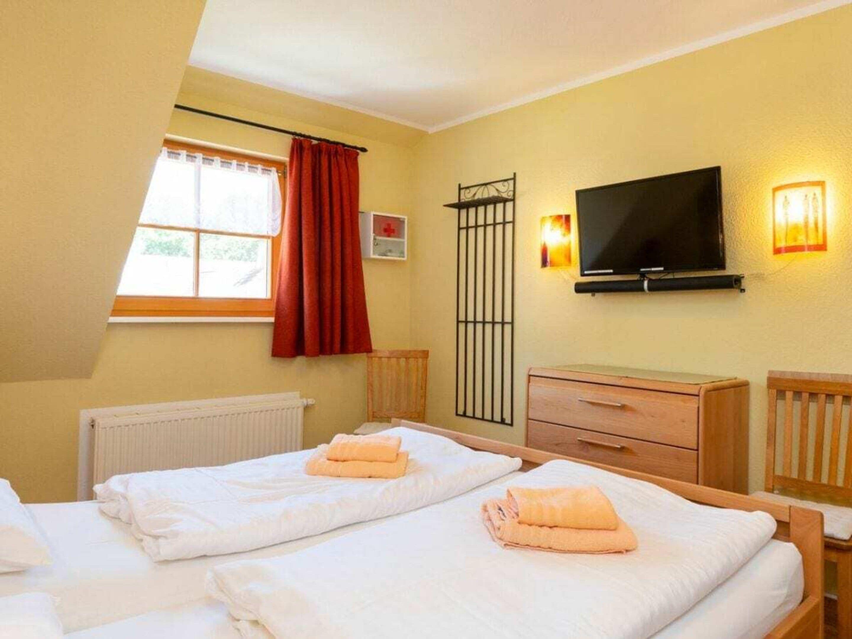 A hotel room with two beds, a television, a window with curtains, a radiator, and a wooden cabinet.