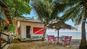 Una cabaña frente a la playa con techo de paja, una pequeña mesa con sillas y vistas al océano.