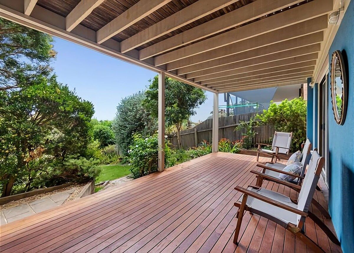 A covered outdoor deck with wooden flooring, two lounge chairs, and a view of a garden with trees and a fence.