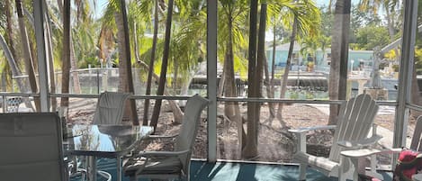 A covered outdoor seating area with a table and chairs, surrounded by palm trees and a view of a marina.